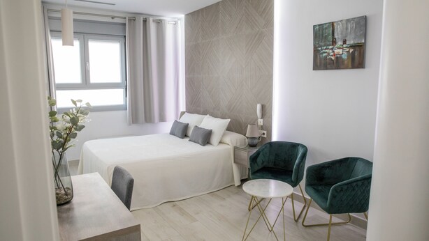 A modern hotel room with a large bed, two green armchairs, a small round table, and a painting on the wall.