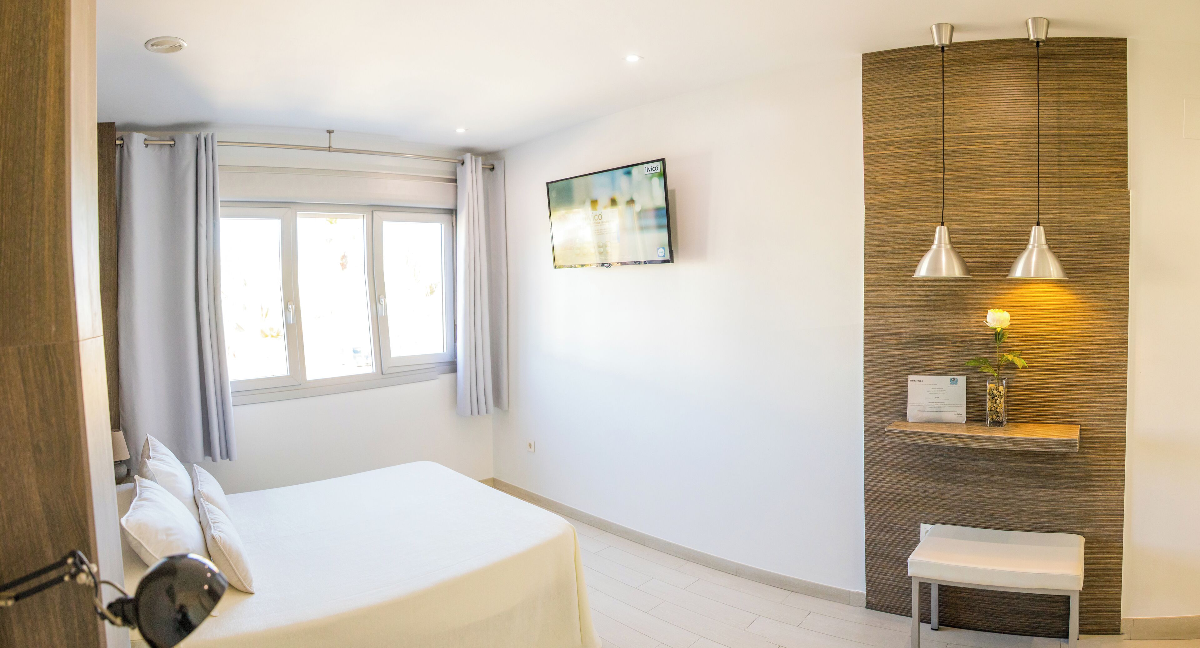 Une chambre d’hôtel comprenant un lit, un bureau, une chaise, une fenêtre avec des rideaux, une étagère fixée au mur avec une plante et un tableau.