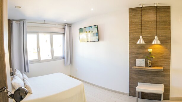 Habitación de hotel con cama, escritorio, silla, ventana con cortinas, estante en la pared con una planta y un cuadro.