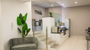 A modern living room with a grey sofa, a large green plant, and a staircase leading to a kitchen area.