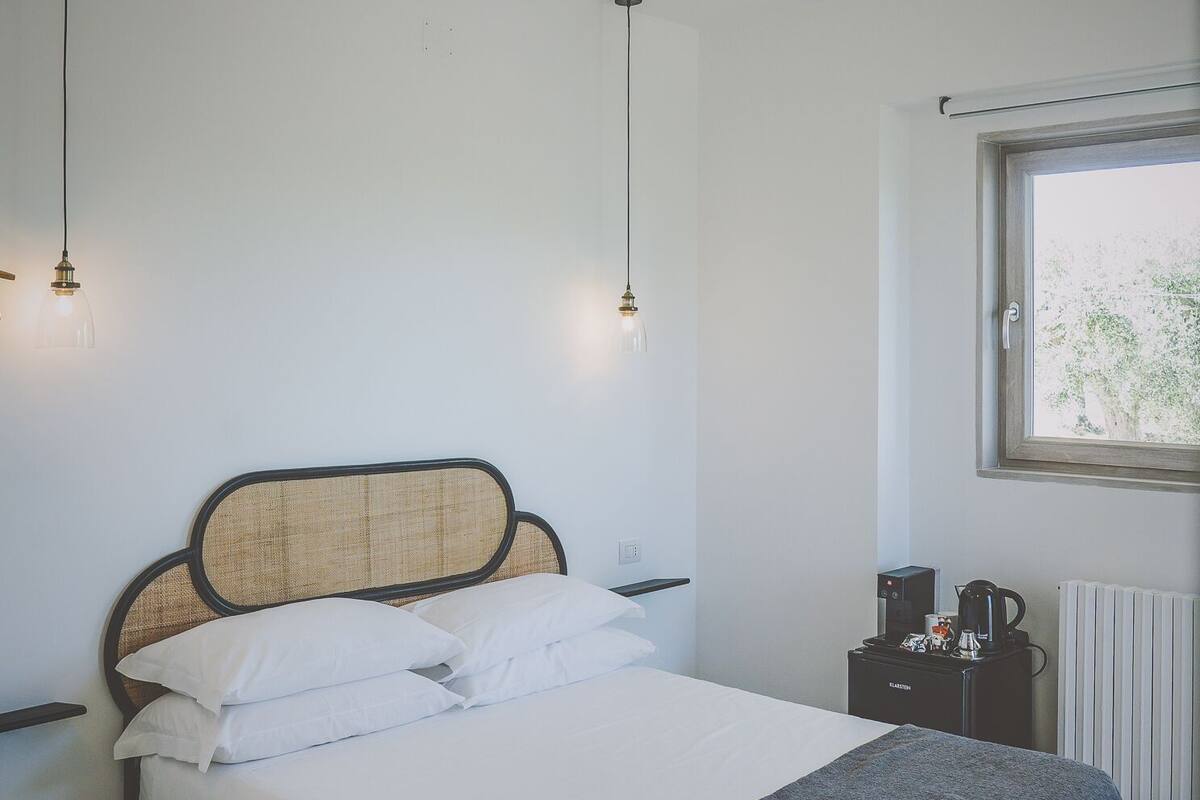 A neatly made bed with white linens and a dark throw blanket, a bedside table with a kettle and a cup, and a window with a view of trees.