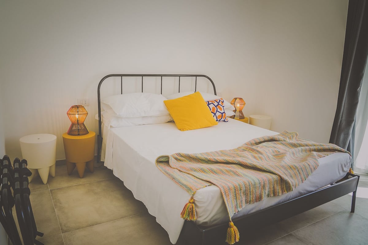 A neatly made bed with white linens and a yellow pillow, flanked by two bedside lamps.