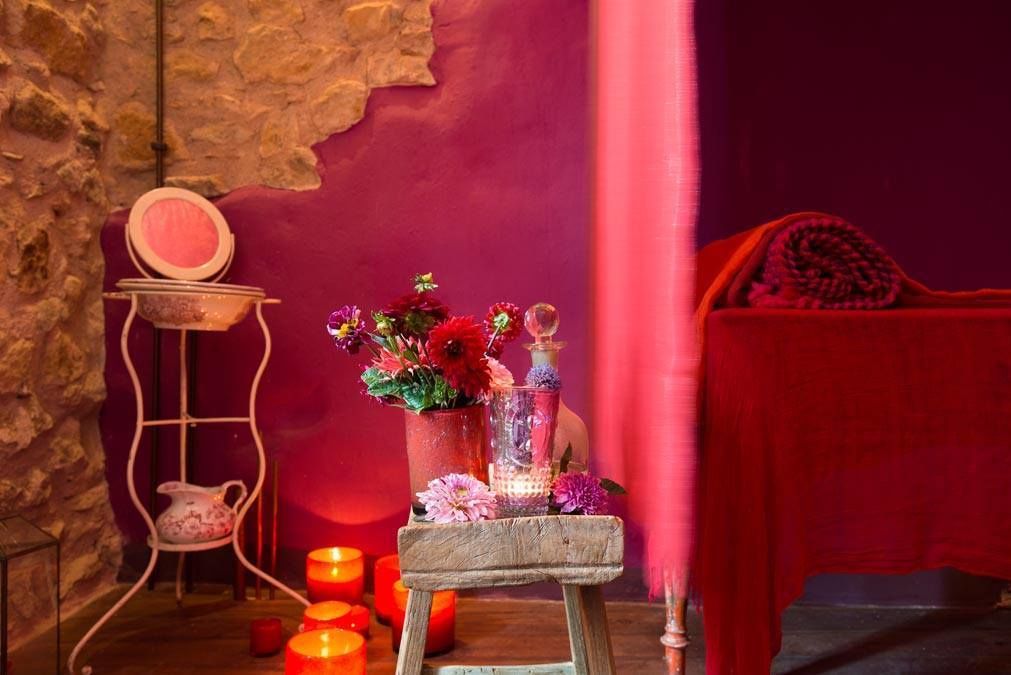 Une pièce avec un mur en pierre rustique, un mur rose, une chaise en bois, une petite table avec des bougies et des fleurs, et un lit rouge.