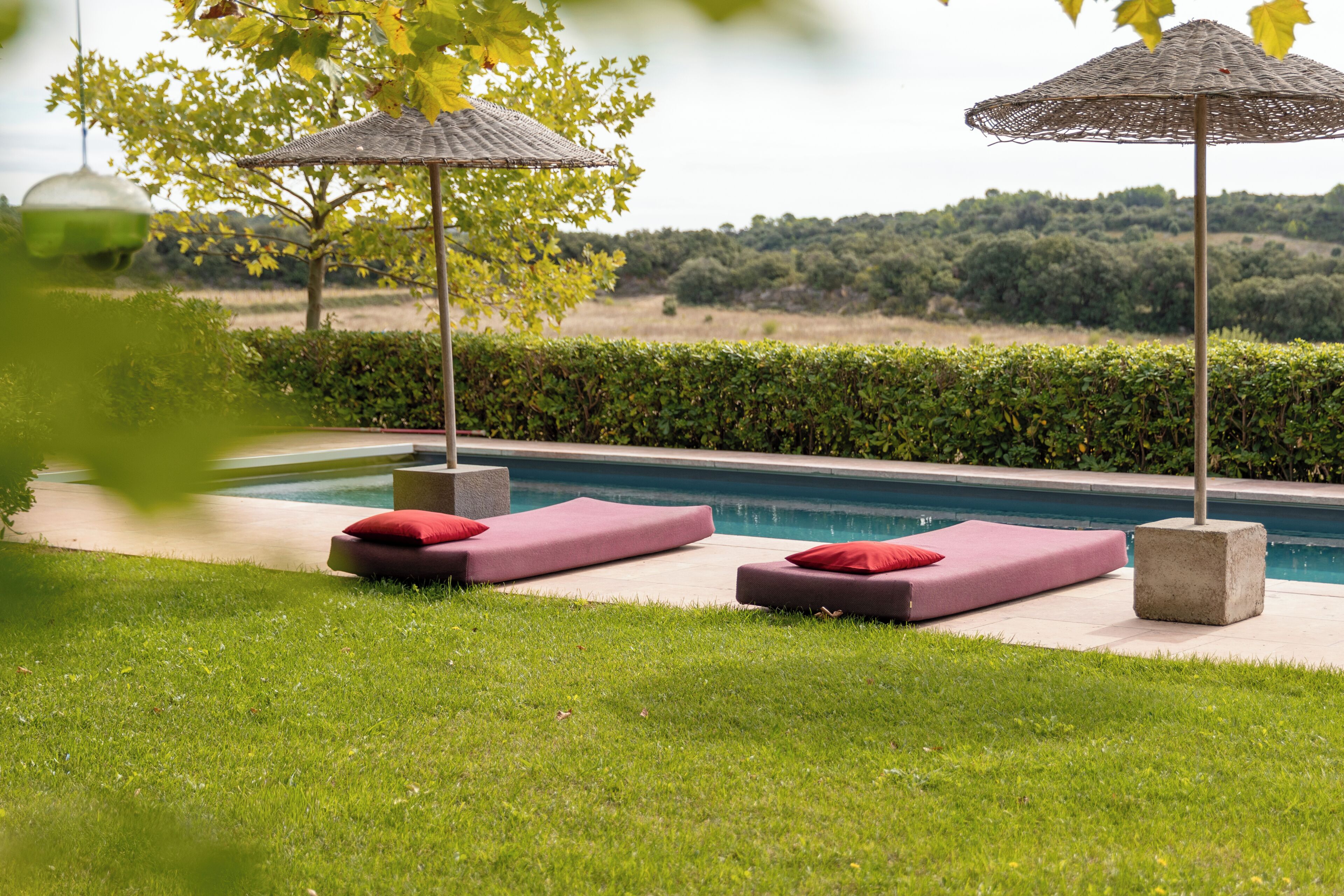 A poolside area with two lounge chairs and umbrellas, a clear view of the pool, and a hedge in the background.