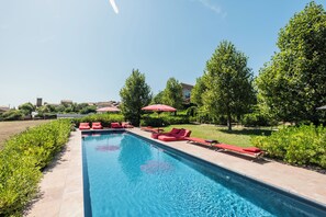 A swimming pool with red lounge chairs and umbrellas, surrounded by greenery and a clear sky. - Village Castigno - Wine hotel & resort (Assignan)