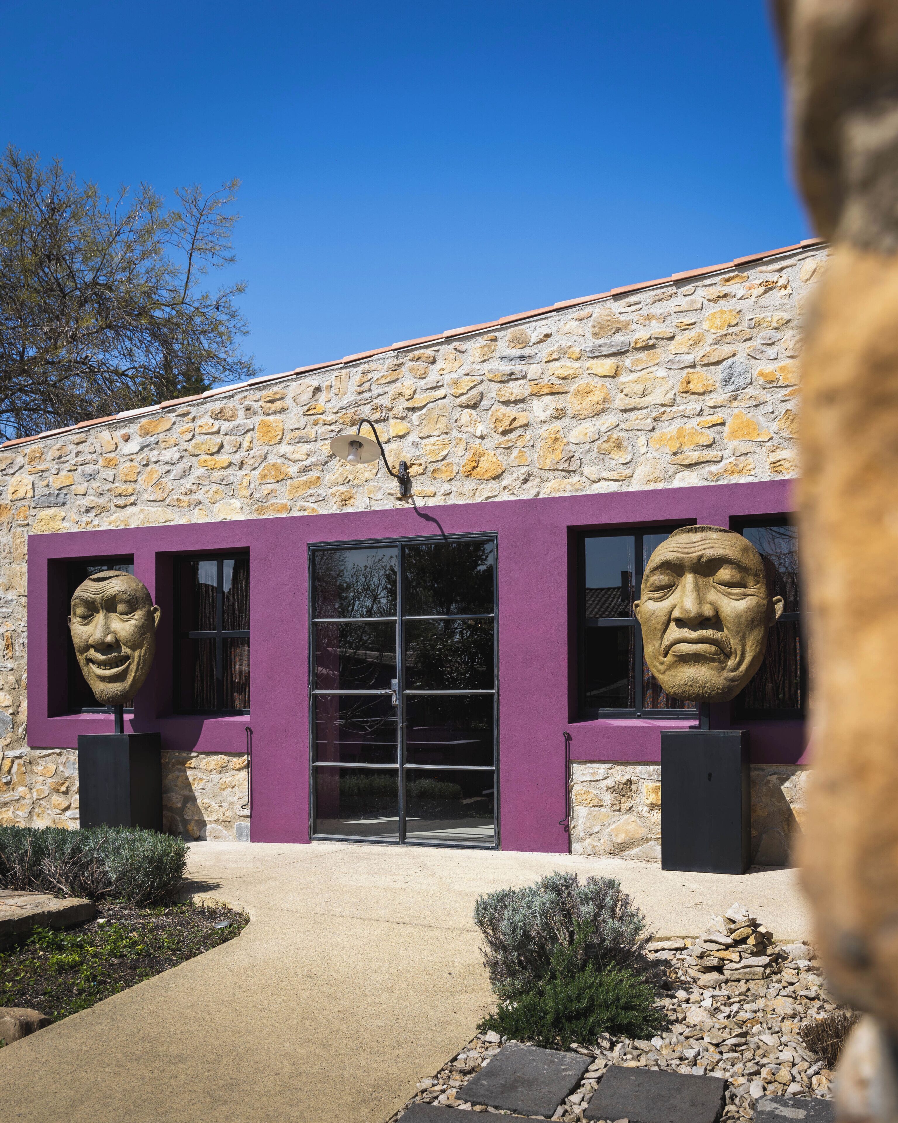 Un bâtiment doté d’une façade en pierre, agrémentée de touches de couleur violette, et de deux grandes sculptures de part et d’autre de l’entrée.