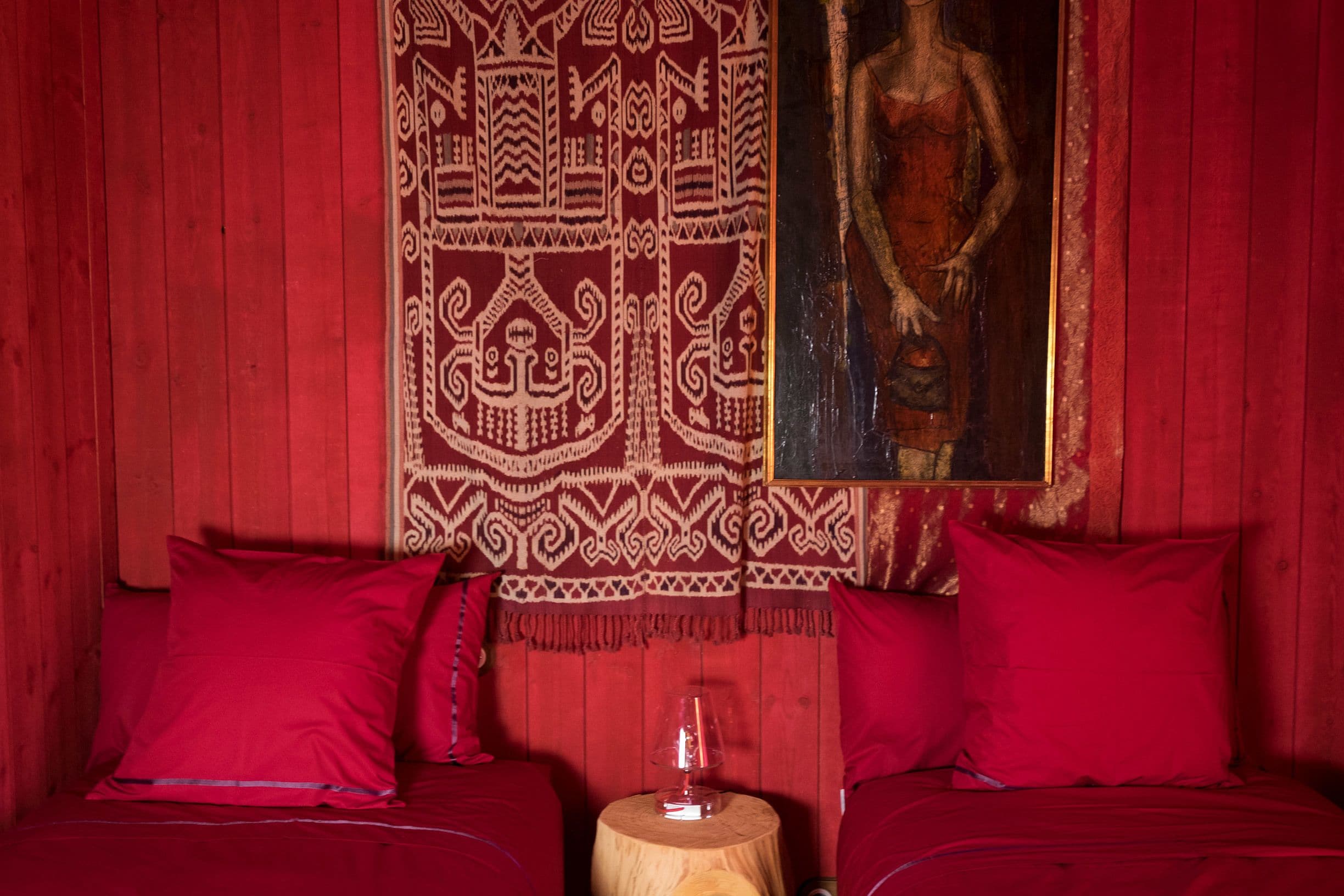 A room with a red wooden wall, a patterned tapestry, and a painting of a woman. There are two red pillows and a small wooden table with a glass object on it.