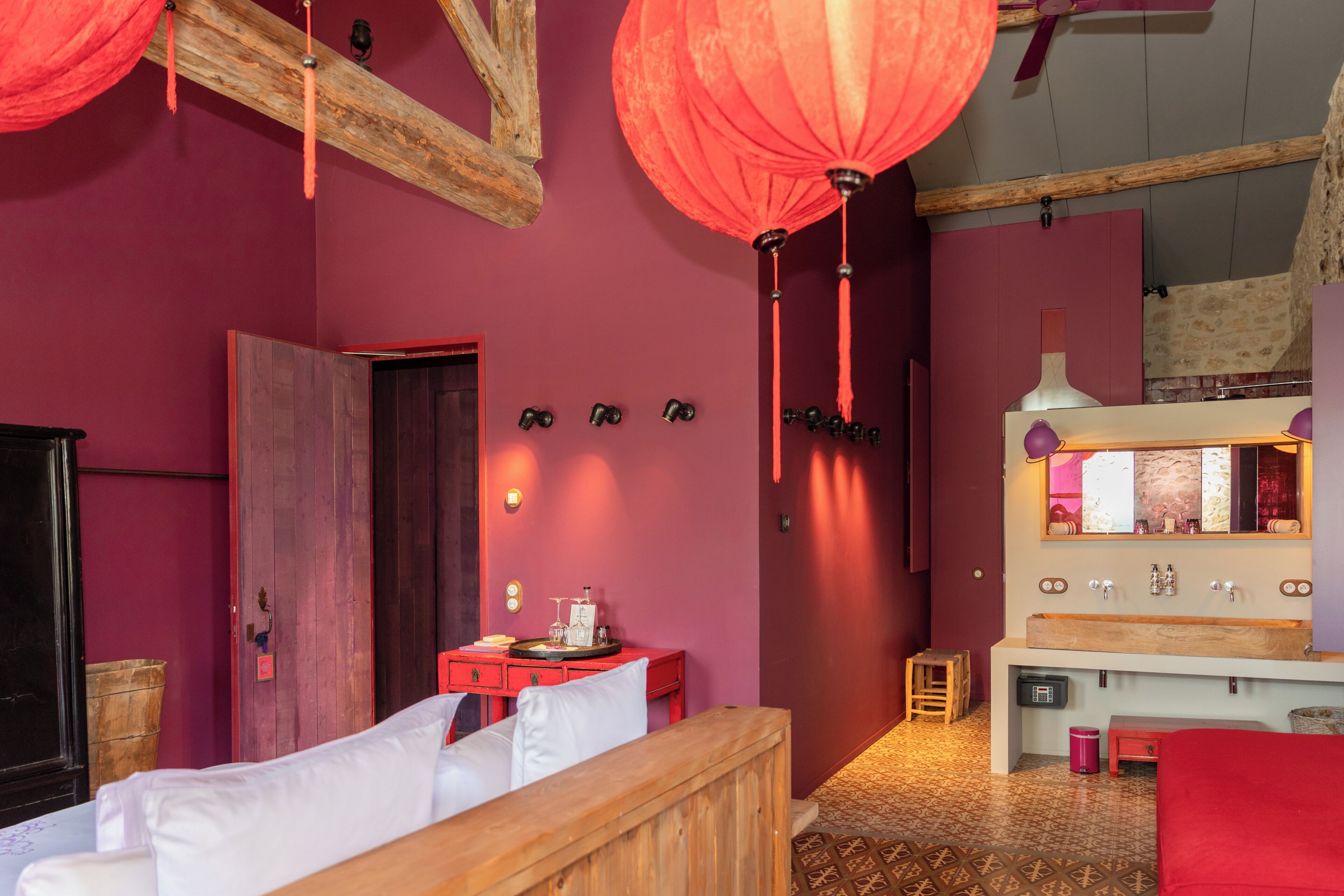 A room with a wooden bed, a red wall, a wooden bench, a small table with a lamp, and a mirror above a sink.