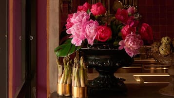 Una habitación con un gran jarrón negro lleno de flores rosas y hojas verdes, un vaso de espárragos y un plato decorativo con alcachofas.