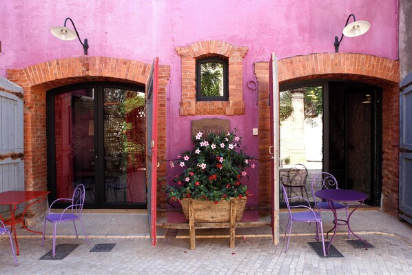 A pink building with brick arches, a wooden planter with flowers, and purple metal chairs and tables. - Village Castigno - Wine hotel & resort (Assignan)