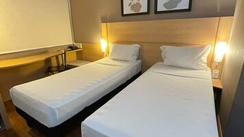 Quarto de hotel com duas camas de solteiro, uma escrivaninha, uma cadeira, televisão e quadros nas paredes.