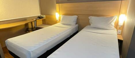 A hotel room with two single beds, a desk, a chair, a TV, and framed artwork on the wall.