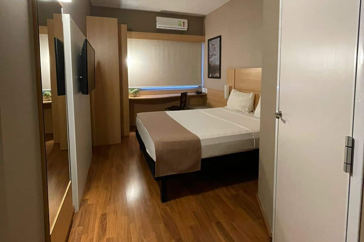 Ett modernt hotellrum med trägolv, en säng med sänggavel, ett skrivbord med stol, en spegel och en väggmonterad luftkonditioneringsanläggning.
