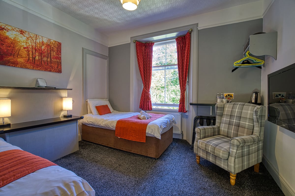A hotel room with a bed, a chair, a window with curtains, a painting on the wall, and a shelf with a lamp.