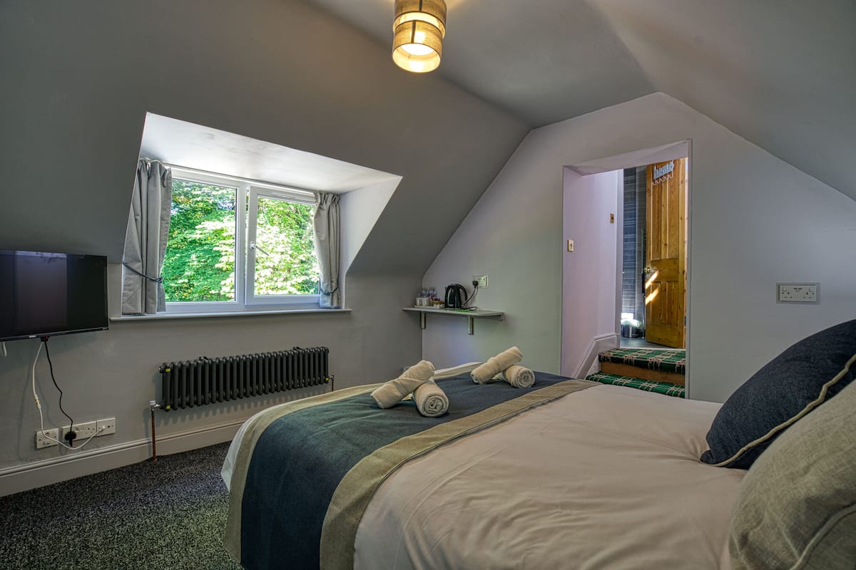 A bedroom with a large bed, a window with curtains, a radiator, a television, and a door leading to another room.