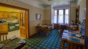 A traditional kitchen with wooden cabinets, a dining area with a green patterned tablecloth, and a window with lace curtains. - Woodlands Bed & Breakfast (Dornoch)