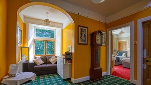 A room with a green patterned carpet, a brown sofa with cushions, a white table with a lamp, and a large wooden clock on the wall. - Woodlands Bed & Breakfast (Dornoch)