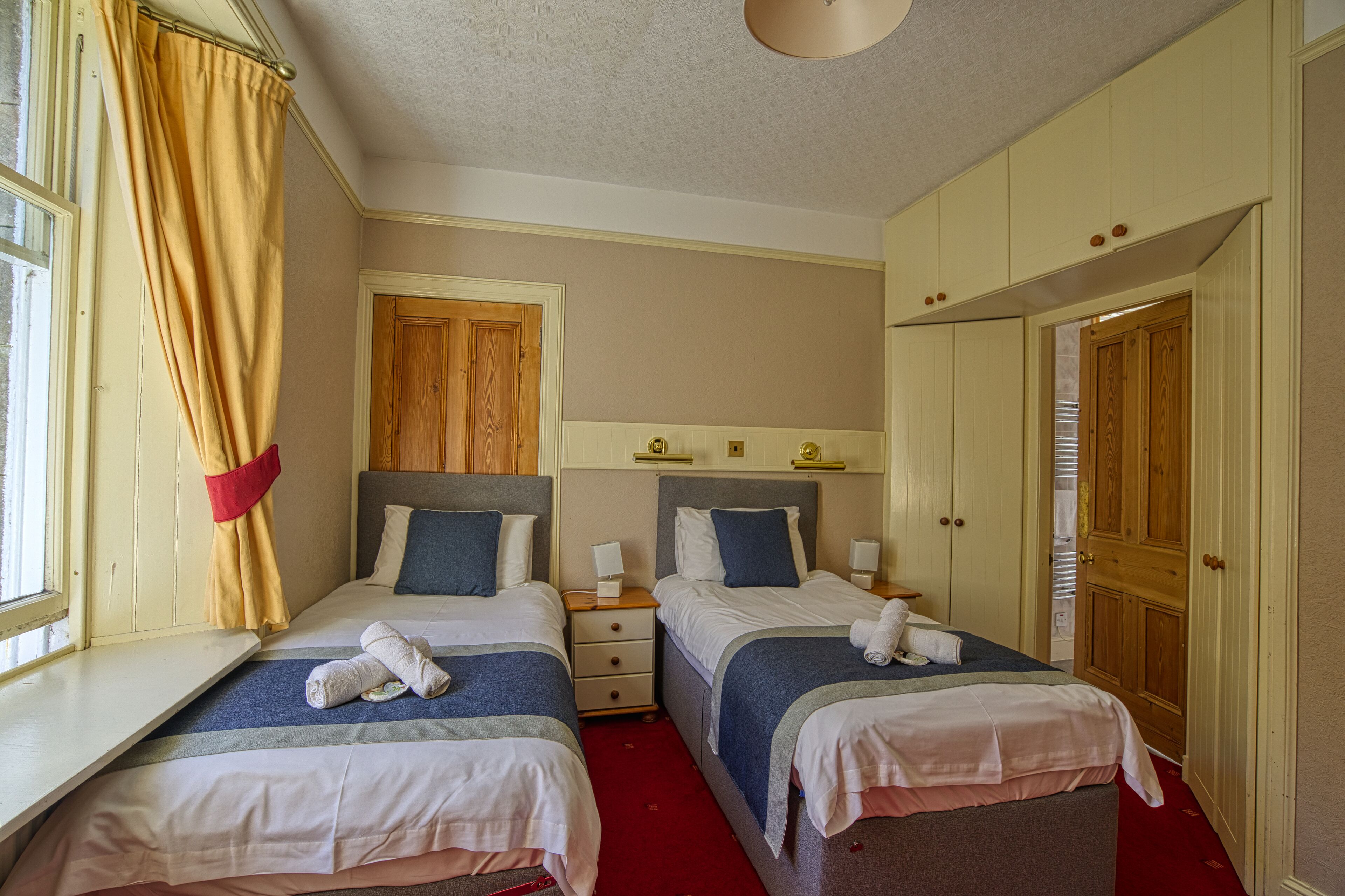 A hotel room with two beds, a wooden door, a window with curtains, and a red carpet.