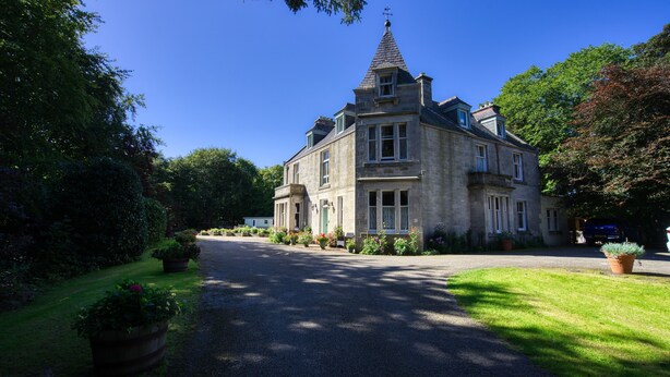 A large stone building with a prominent tower, surrounded by well-maintained gardens and a paved driveway. - Woodlands Bed & Breakfast (Dornoch)