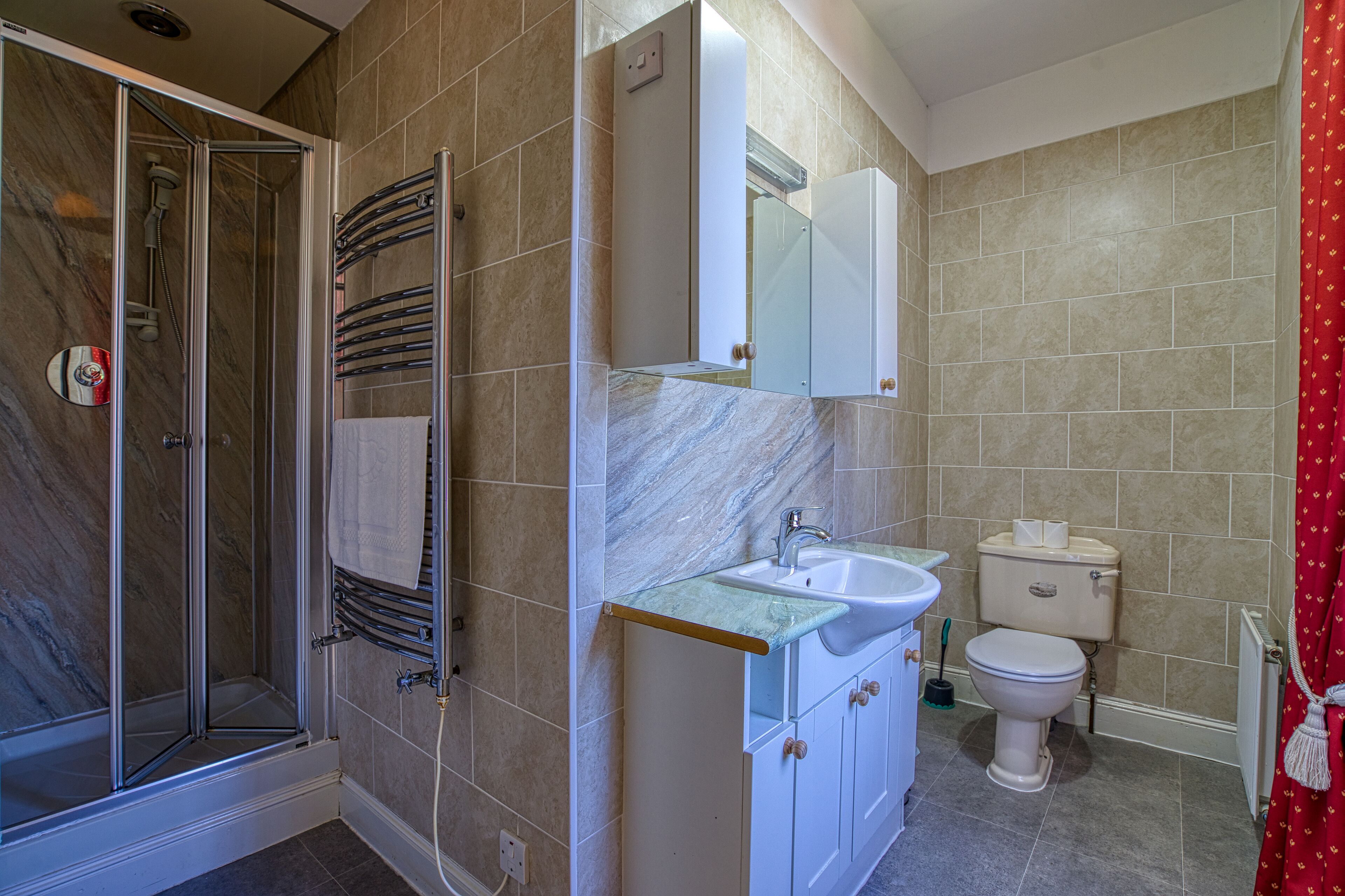 A bathroom with a glass shower enclosure, a sink with a marble countertop, a toilet, and a red shower curtain.