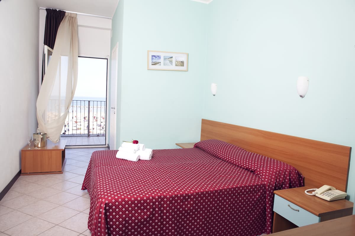 Una habitación de hotel con una cama, una mesita de noche con un teléfono, otra mesita más pequeña con una lámpara, una cortina y un cuadro en la pared.