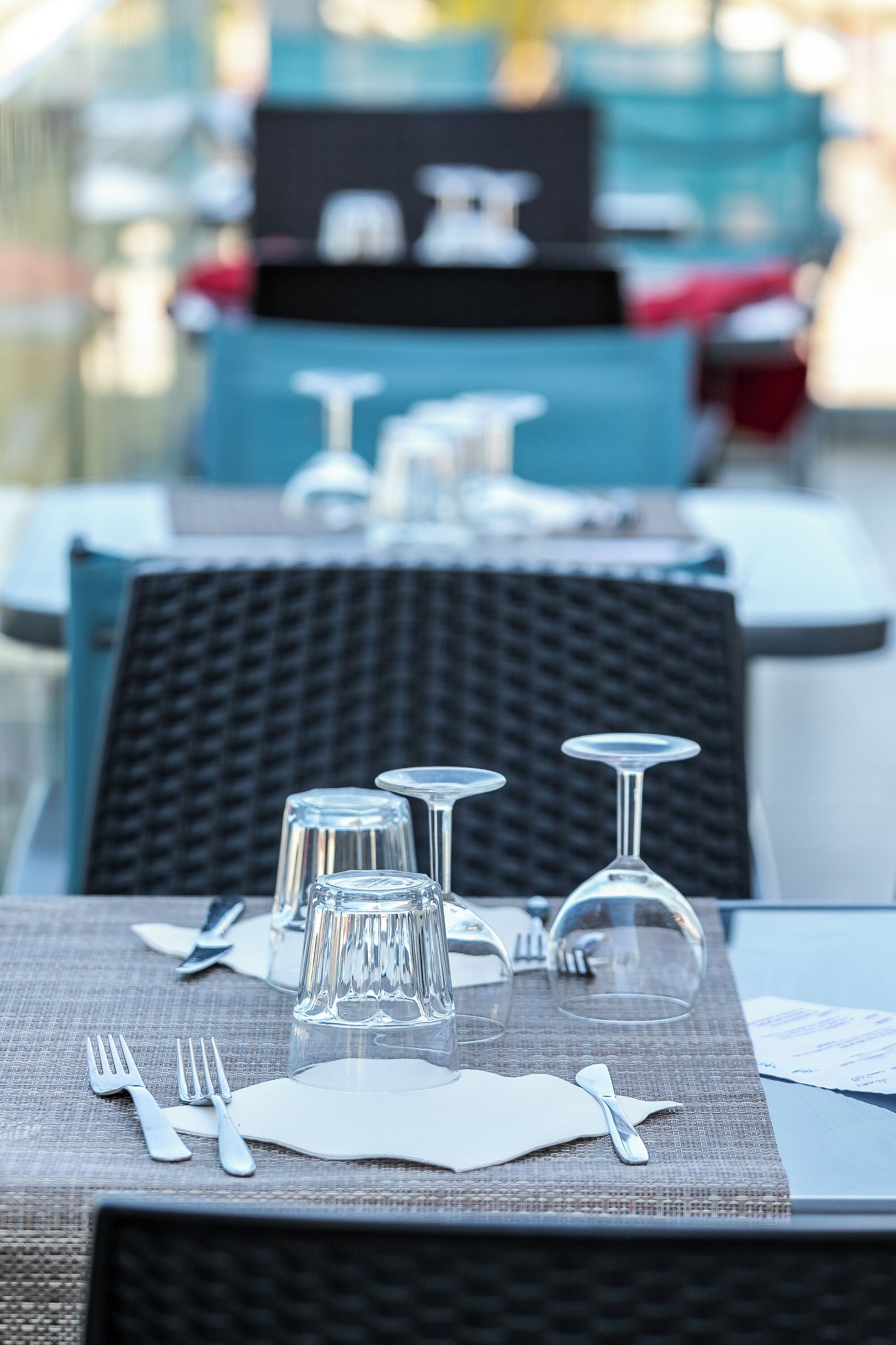Una mesa de comedor dispuesta con cubiertos y cristalería, con una silla de mimbre y un cojín azul al fondo.