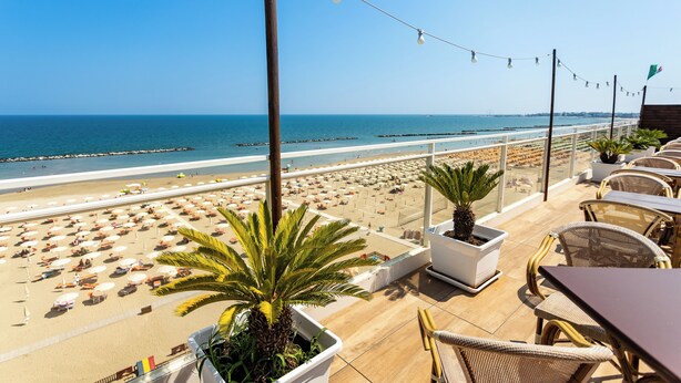 A beachside terrace with outdoor seating, a view of the beach, and a palm tree. - Hotel Blumen (Rimini)