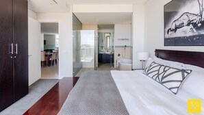 A modern hotel room with a large bed, a dark wooden wardrobe, a bathroom with a shower, and a dining area visible through an open door. - 606 Juliette B (Cape Town)