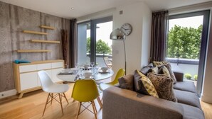 A modern living room with a sofa, a dining table, and chairs. There is a large window with a view of trees.