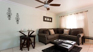 A living room with a sectional sofa, a coffee table, a ceiling fan, and a dining area with a small table and stools.