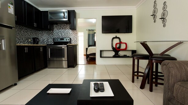 A modern kitchen with dark cabinets, a microwave, and a tiled backsplash. There is a dining area with a glass table and two bar stools. A TV is mounted on the wall, and a flat-screen TV is on a stand.