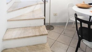 A staircase with wooden steps leading to a higher floor, a white wall with a colorful abstract painting, a round table with chairs, and a floor lamp. - Luna (Florence)
