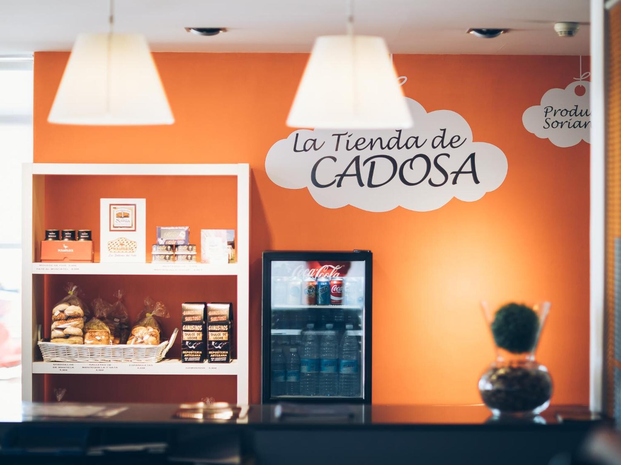 A bakery counter with a sign reading "La Tienda de CADOSA" and a refrigerator stocked with beverages.