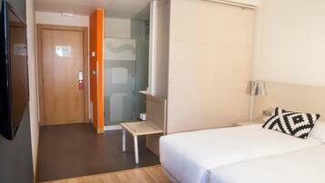 A hotel room with a bed, a bedside table, a lamp, a TV, and a shower area.