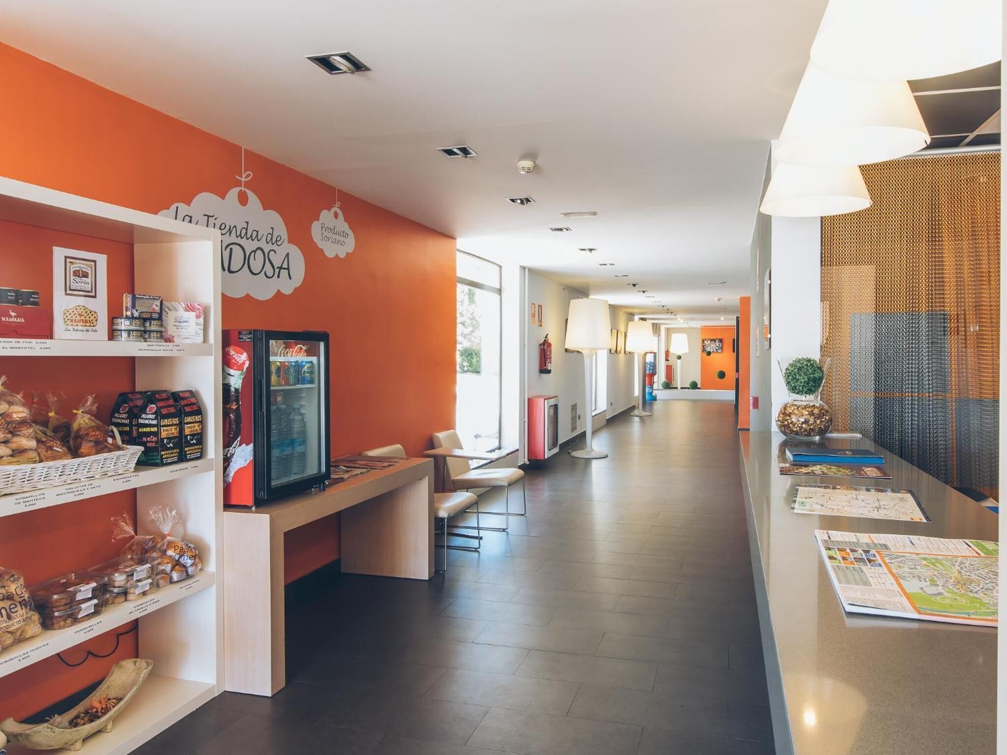 Ein Korridor mit Regalen links, auf denen Snacks und eine Coca-Cola-Kühlbox ausgestellt sind, einer Bank und einem Tisch mit Zeitschriften.