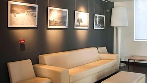 A modern living room with a beige sofa, a matching ottoman, and a coffee table. There are framed pictures on the wall and a floor lamp. - Hotel Cadosa (Soria)