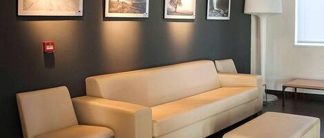 Un salón moderno con un sofá color beige, un reposapiés a juego y una mesa de centro. Hay cuadros enmarcados en la pared y una lámpara de pie.