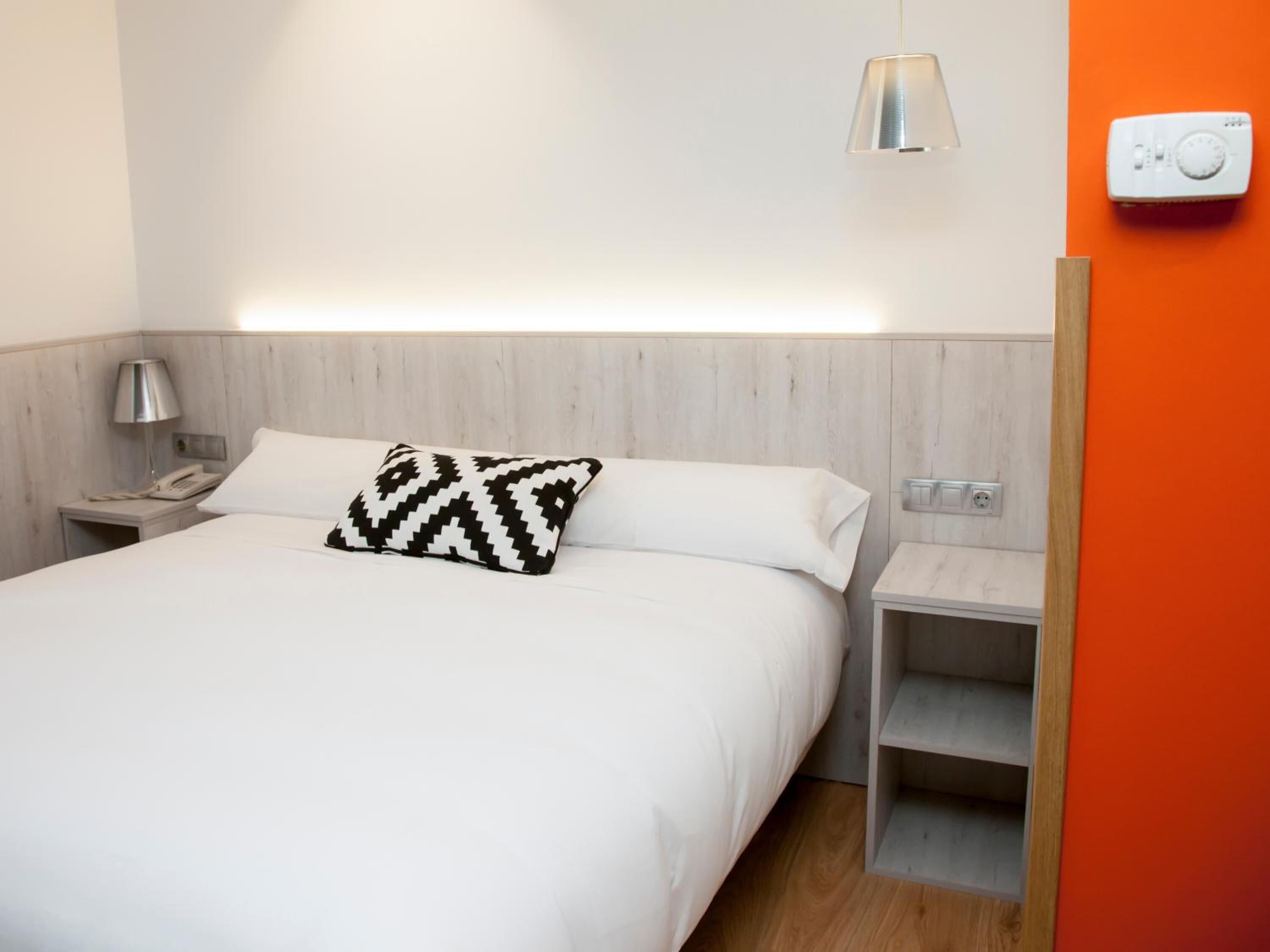 Habitación de hotel con una cama, una mesita de noche con lámpara, un termostato y un estante pequeño.
