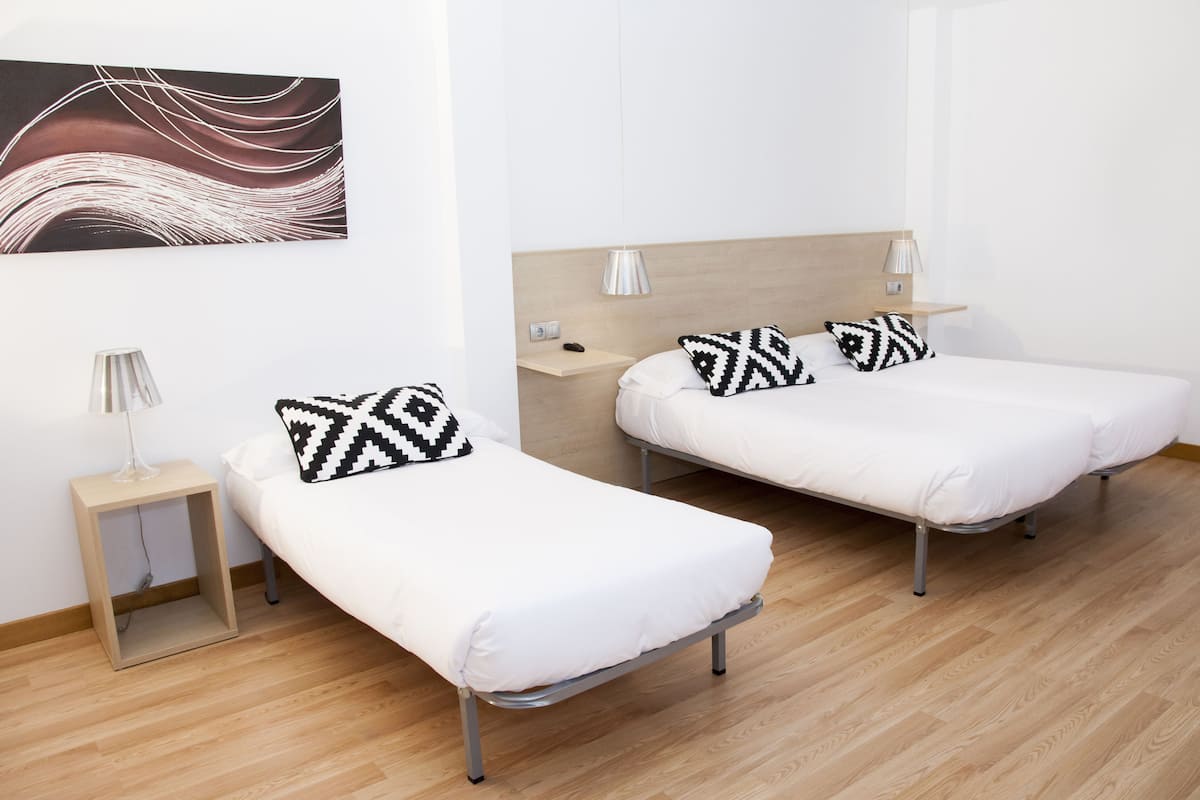 Habitación de hotel con dos camas individuales, una mesita de noche con lámpara, estante en la pared y un cuadro abstracto moderno.