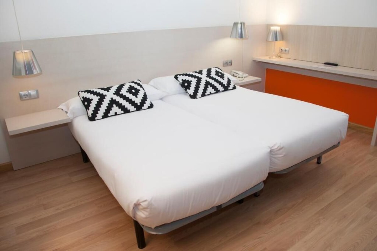 A double bed with white bedding and black and white patterned pillows, two bedside tables with lamps, and an orange accent wall.