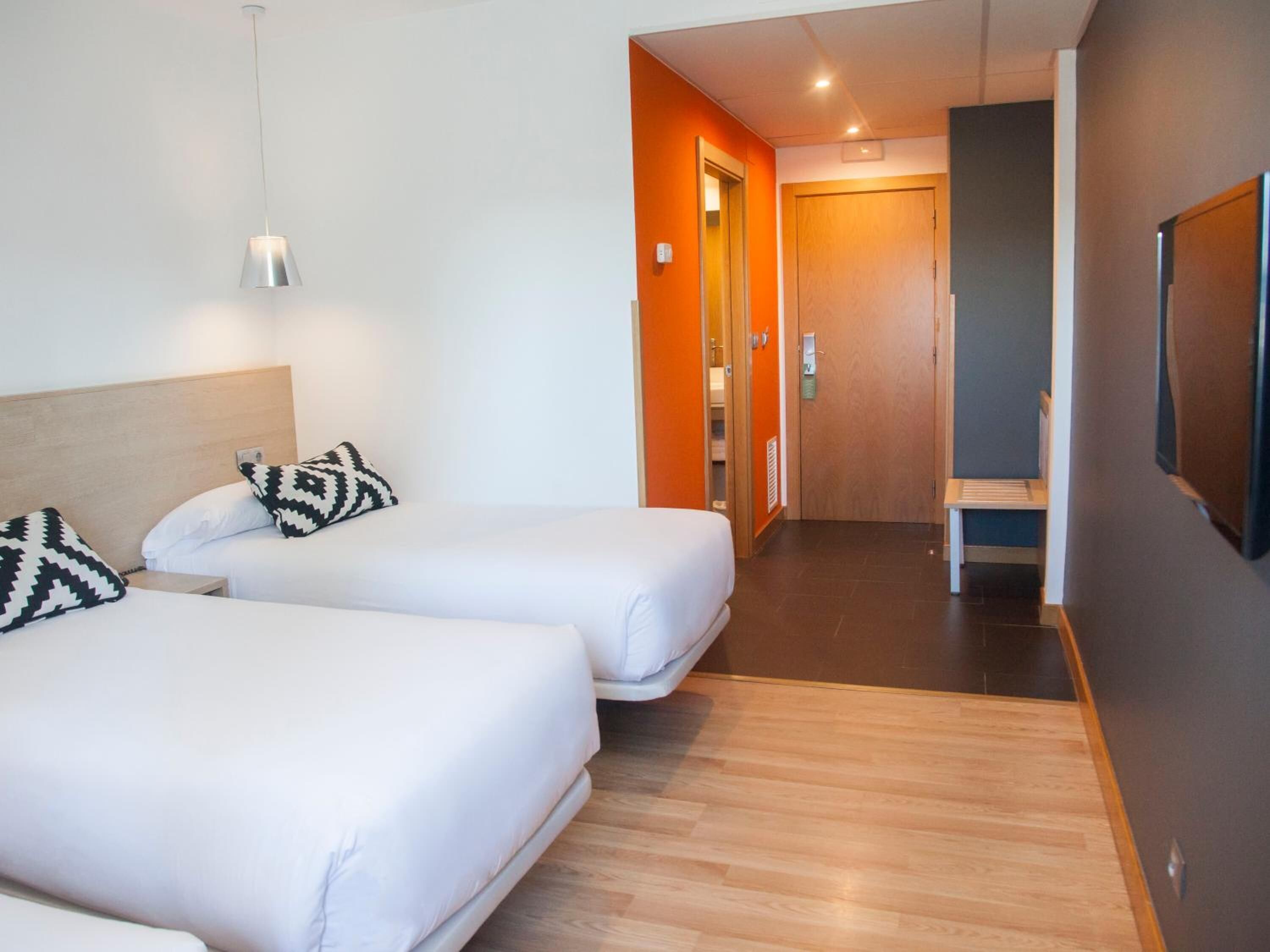 A hotel room with two beds, a wooden headboard, a hanging light fixture, and a door leading to a bathroom.