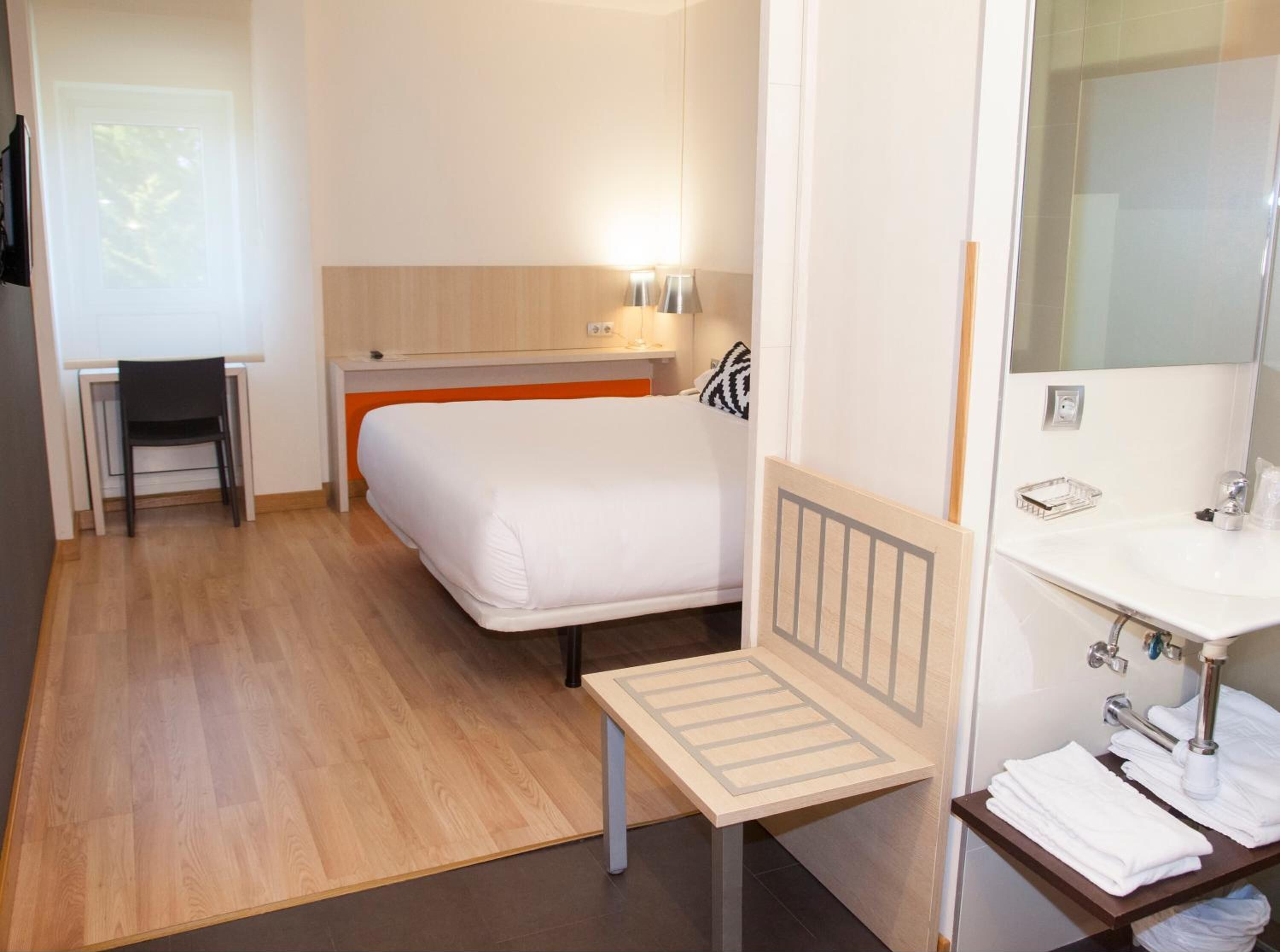 A hotel room with a bed, a desk, a chair, a small table, a bathroom with a sink and mirror, and a window with a view.