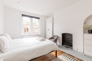 A bedroom with a large bed, a chair, a fireplace, and a view of buildings outside.