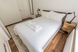 A neatly made bed with white linens and folded towels, a bedside table with a lamp, and a wooden floor.