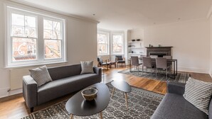 A modern living room with a grey sofa, black coffee table, and patterned rug. There are two windows with a city view, a fireplace, and a dining area with chairs. - Eson2 - Charming 3 Bedroom Flat in Chelsea (London)