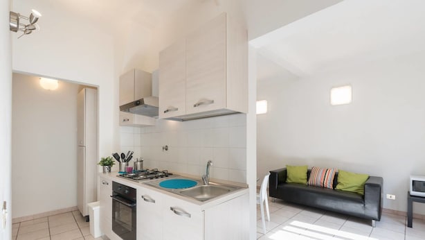 Una cocina moderna con armarios blancos, un lavamanos azul y una estufa a gas. En la sala de estar, hay un sofá de cuero negro con almohadones coloridos.