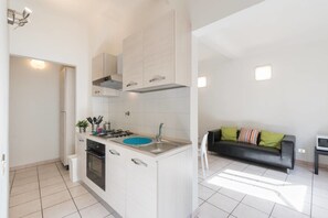 Una cocina moderna con armarios blancos, un lavamanos azul y una estufa a gas. En la sala de estar, hay un sofá de cuero negro con almohadones coloridos.