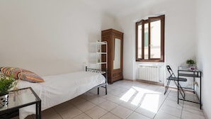 Una habitación pequeña y bien iluminada con una cama individual, un armario de madera, un radiador, una ventana y una pequeña mesa con una silla.