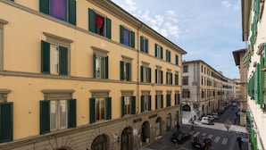 Un hotel con fachada amarilla, contraventanas verdes y vista a la calle con coches aparcados y peatones.