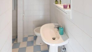 Un baño con lavamanos blanco, un espejo y un inodoro. Hay repisas con artículos de aseo y una pequeña planta.